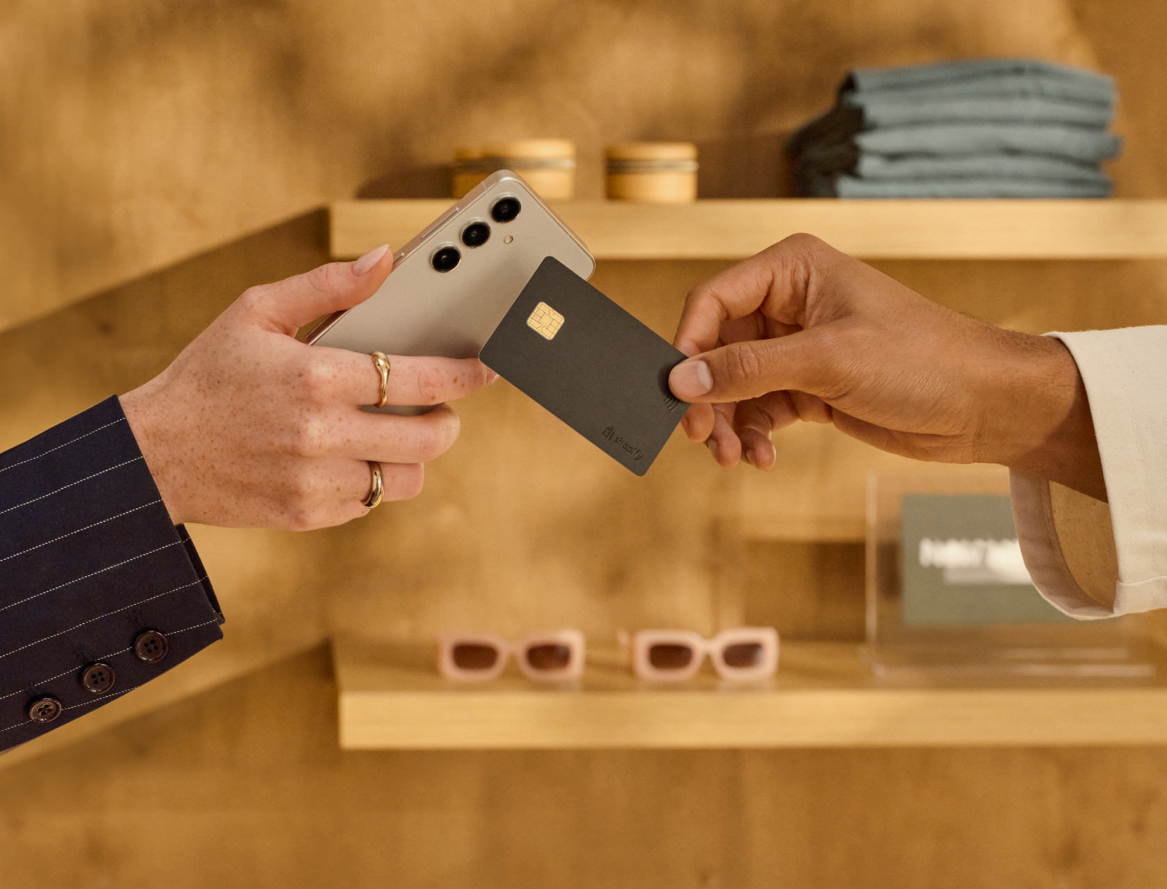 Close-up of a customer using their credit card to make a purchase with Tap to Pay on Android in a large retail environment. The sales associate holds the Android smartphone, while a customer holds their credit card to tap. The background shows a warmly lit store with neatly stocked shelves.