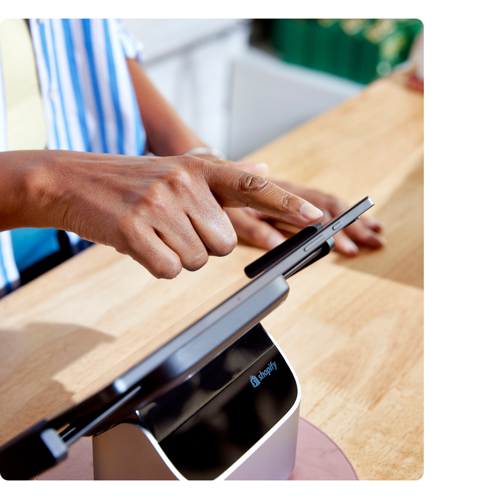 Een close-up van een kassa waar een medewerker met een blauw-wit gestreept overhemd op een POS Tablet typt. De tablet en standaard staan ​​op een toonbank van licht hout. Over de afbeelding heen is een Shopify POS UI-scherm te zien met een betalingsoverzicht.