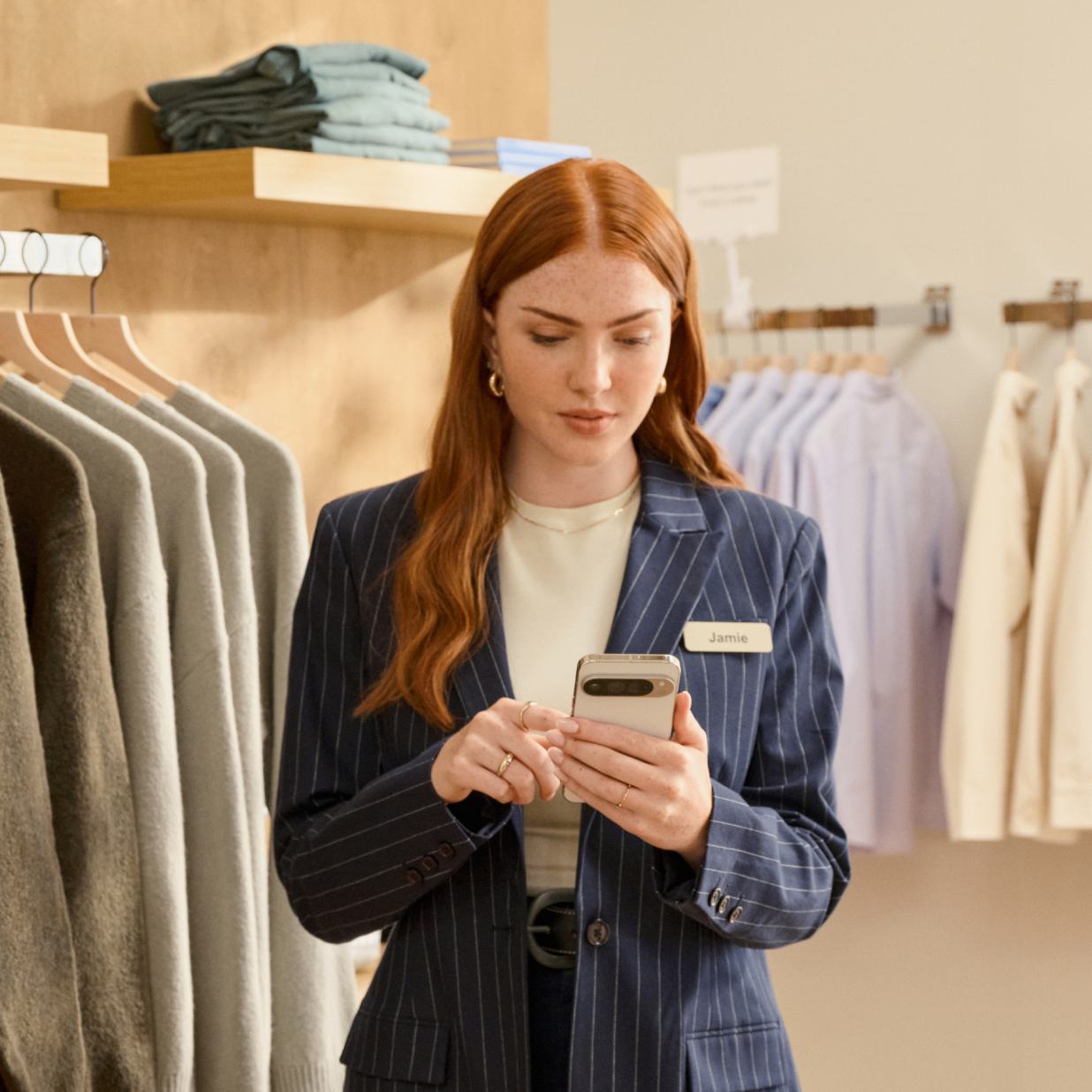 A sales associate with red hair and navy pinstriped blazer, is focused on an Android device in a clothing store. They stand near a rack of clothes featuring a variety of colors including mustard, black, and grey. The store's interior is warmly lit and showcases neatly organized clothing and accessories on wooden shelves and hangers.