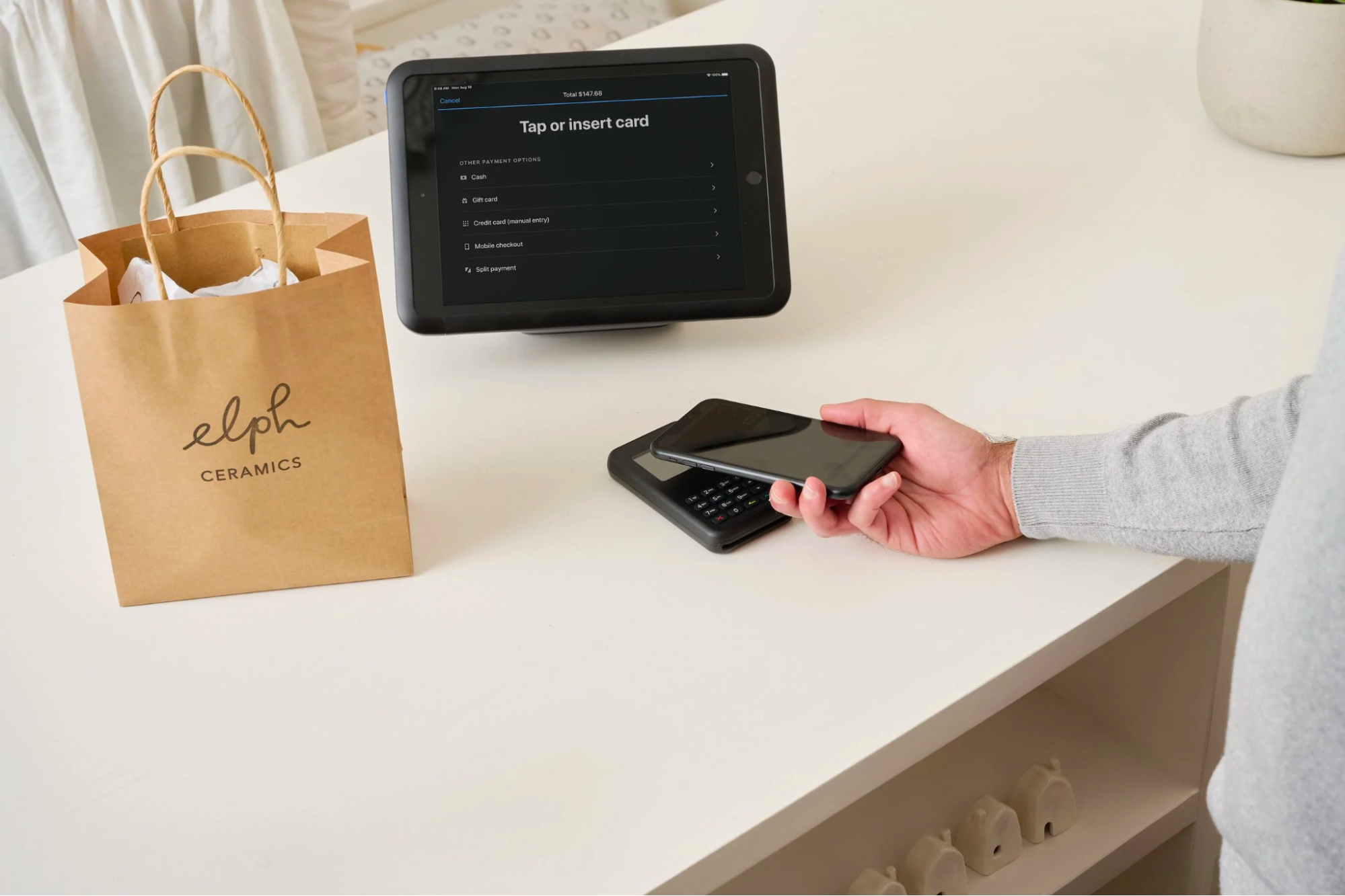 Counter with an elph ceramics bag on it, a Shopify POS nearby, and a hand holding a smartphone.