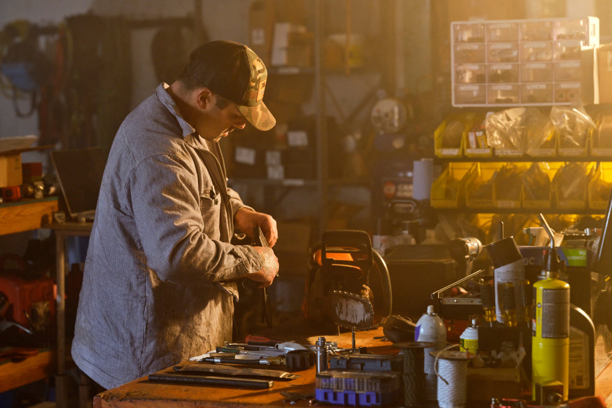 The Shop founder Boe Brodhun is wearing a dark hat and a zip-up shirt, working on a chainsaw at a cluttered workbench in a dark workshop. Various tools and equipment are spread across the bench, and shelves filled with organized boxes and containers are in the background.