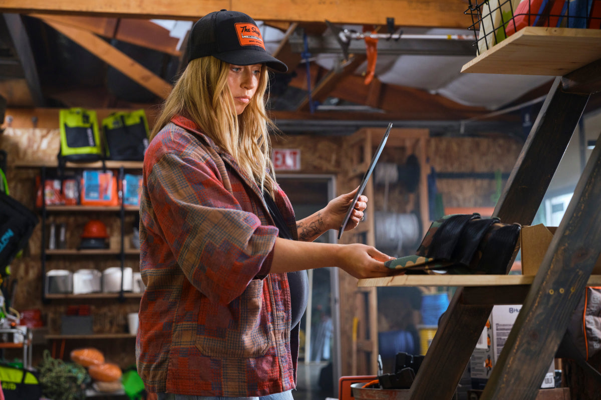 Kennedy Brodhun is wearing a black hat and red plaid shirt inside a workshop. The workshop is filled with various supplies and equipment. She is holding a tablet in her left hand and arranging hats with her right hand.