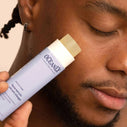A close up photo of a man holding a purple beauty product in front of his face.