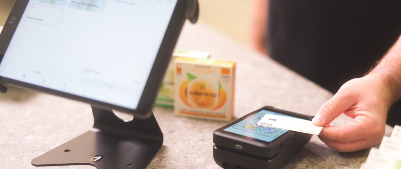 A customer taps a Visa card on a Shopify POS payment terminal at a retail counter, with a tablet screen and product boxes in the background.